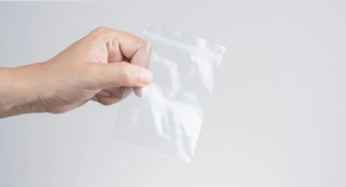 A male's hand holding a small clear plastic zip lock bag against a white wall.